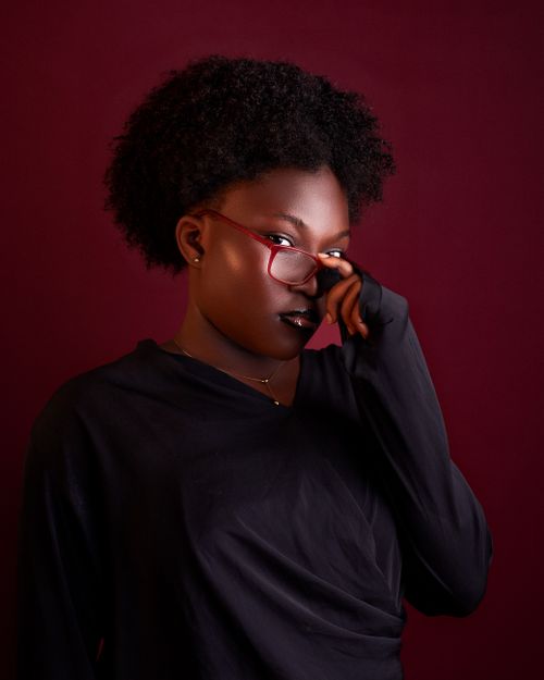 Free Captivating portrait of an adult with red glasses, dark attire against a rich maroon background. Stock Photo
