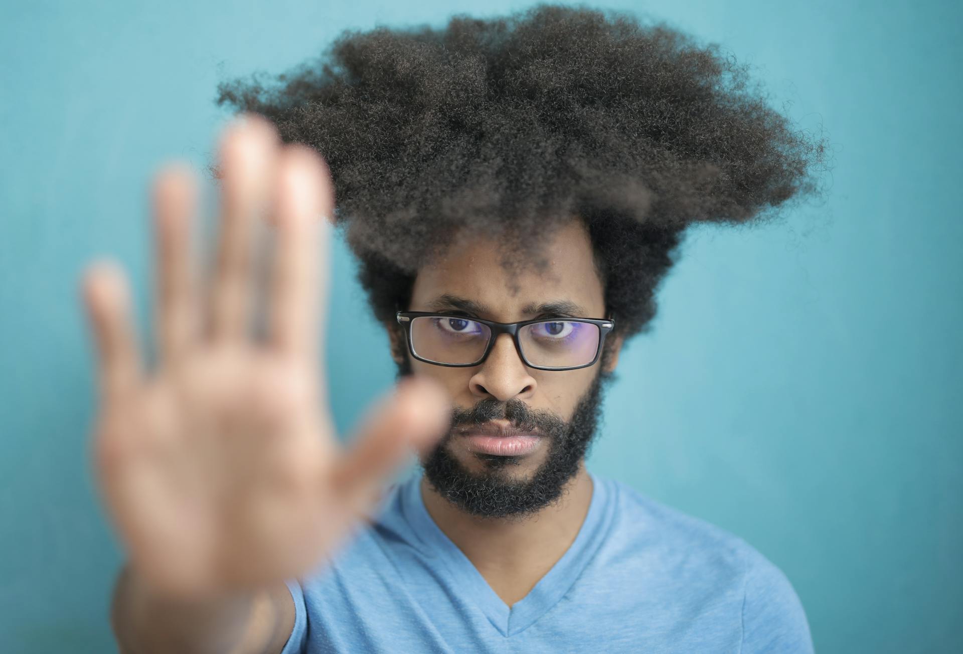 Portrait of a man with afro hair and glasses, making a stop gesture against a blue background.