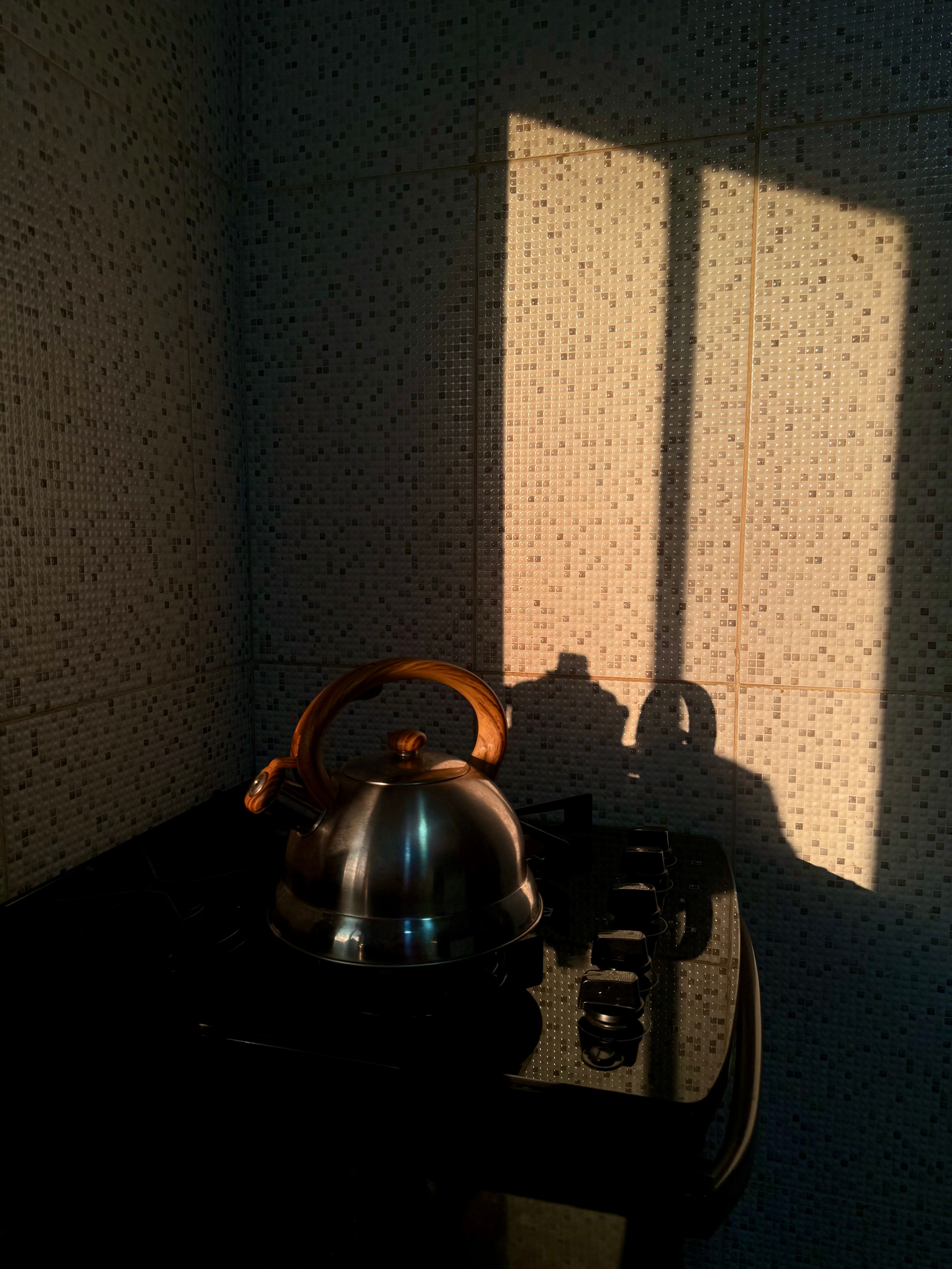 A stainless steel kettle on a stovetop with sunlight casting shadows on the wall.