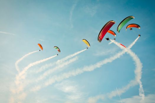 A group of paragliders creates colorful trails against a clear blue sky, displaying aerial skill.