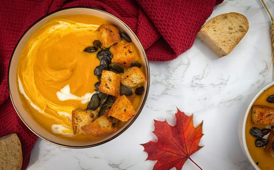 Orange pumpkin soup with cream swirl, sourdough croutons and dark pumpkin seeds beside a maple leaf