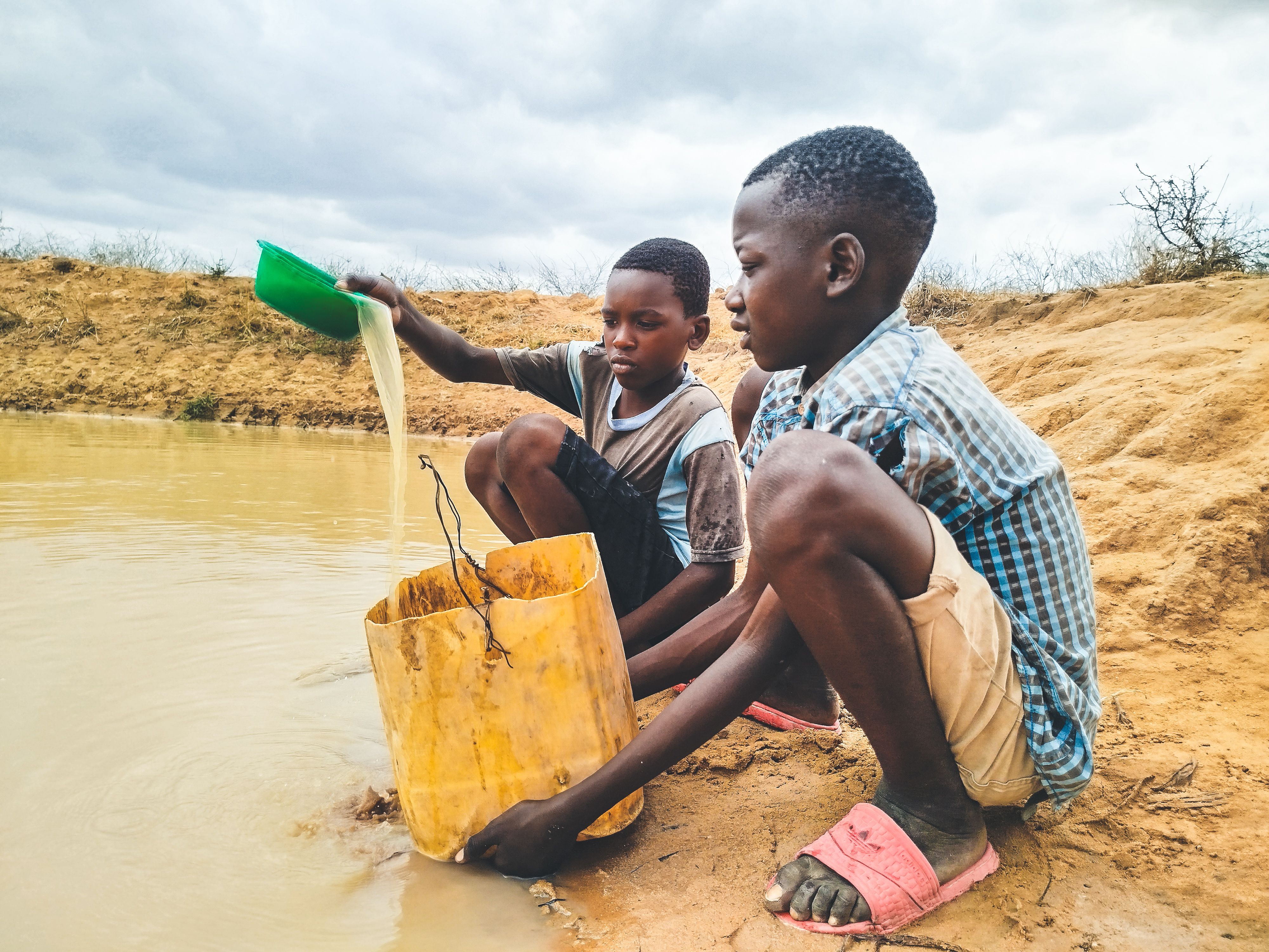Two boys are collecting water from a muddy pond in a rural setting.