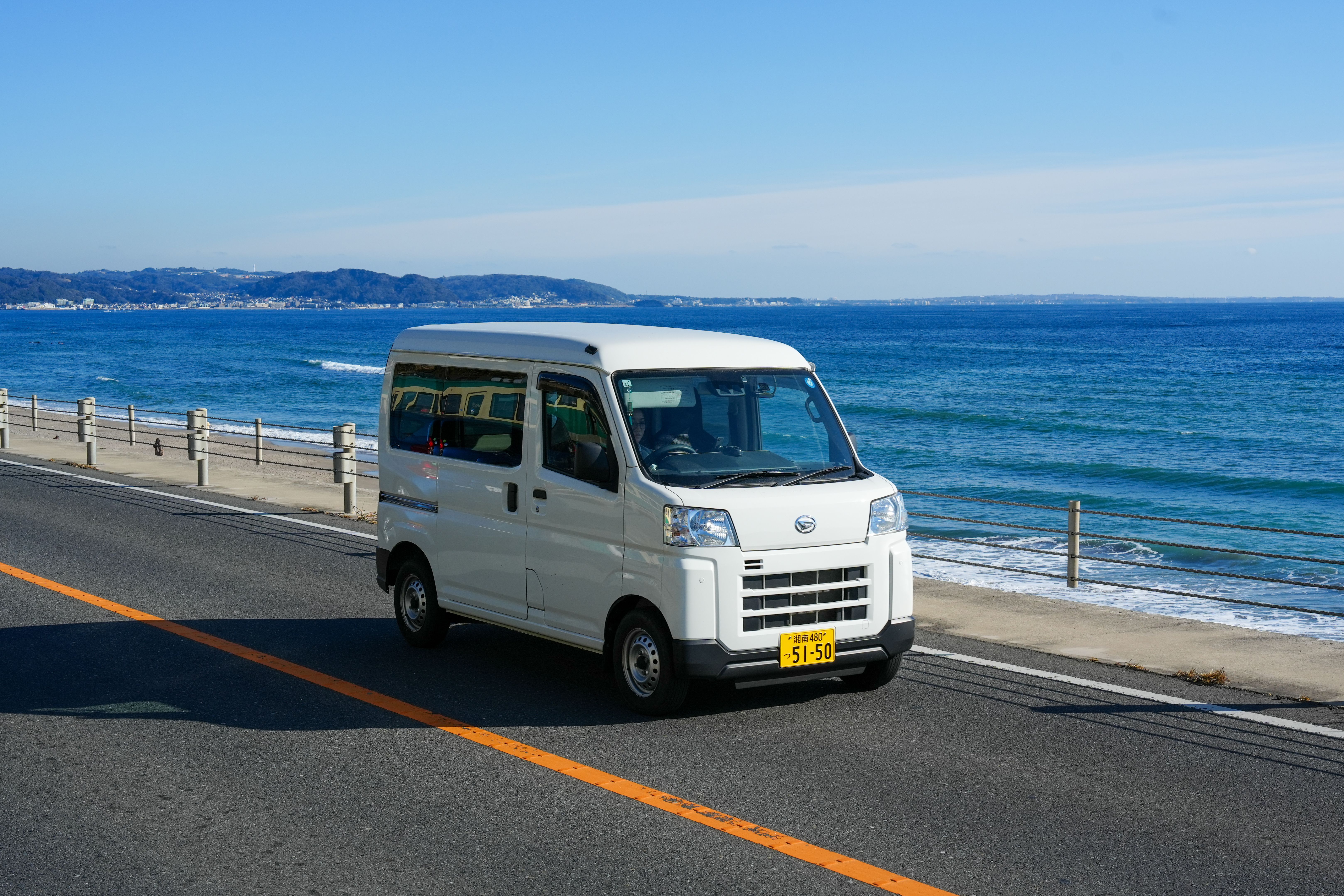神奈川県鎌倉市の海岸沿いの道を走る白いバン。海が見える景色。