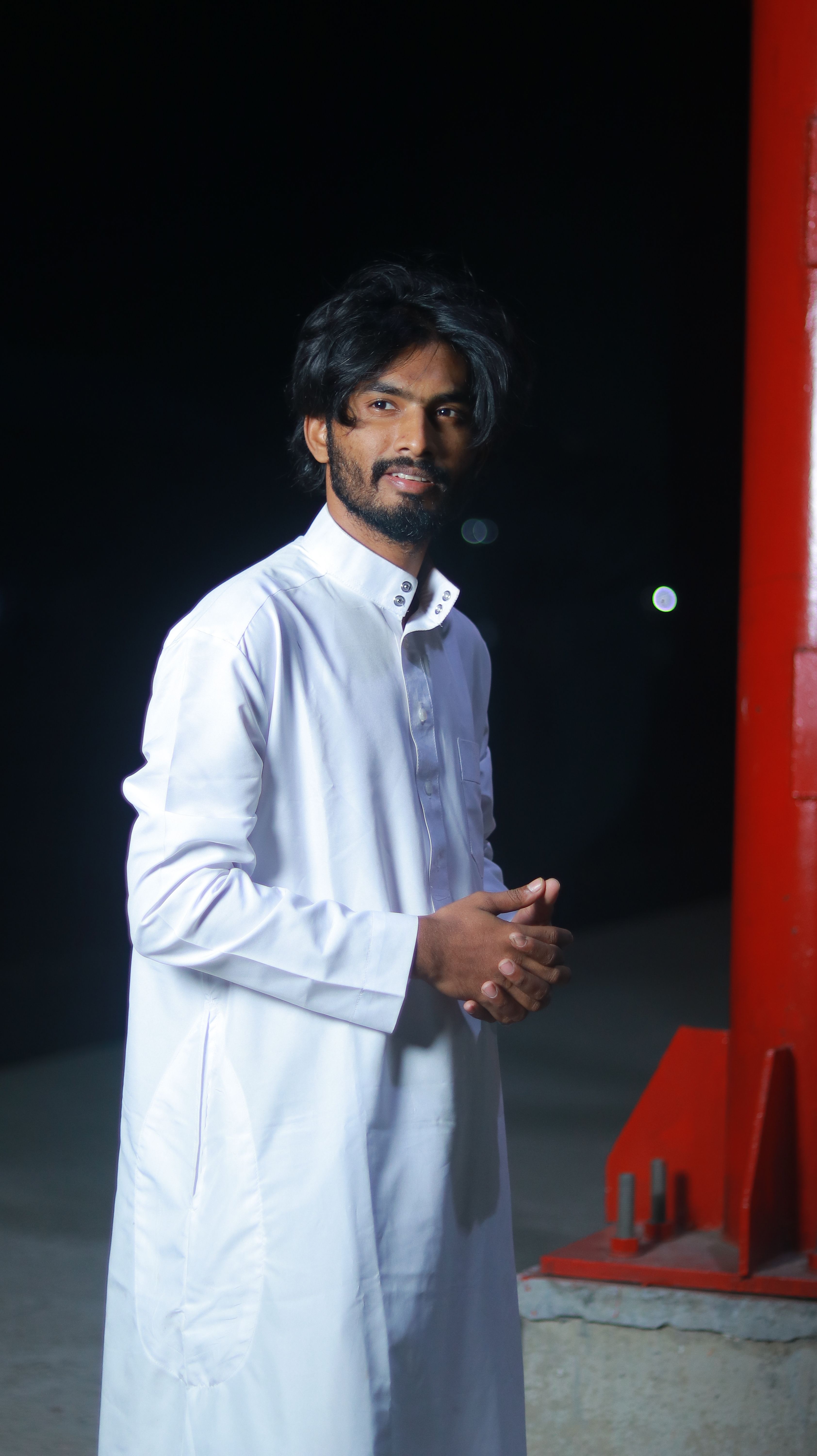 Portrait of a man in a white thobe standing by a red pillar at night, outdoors.