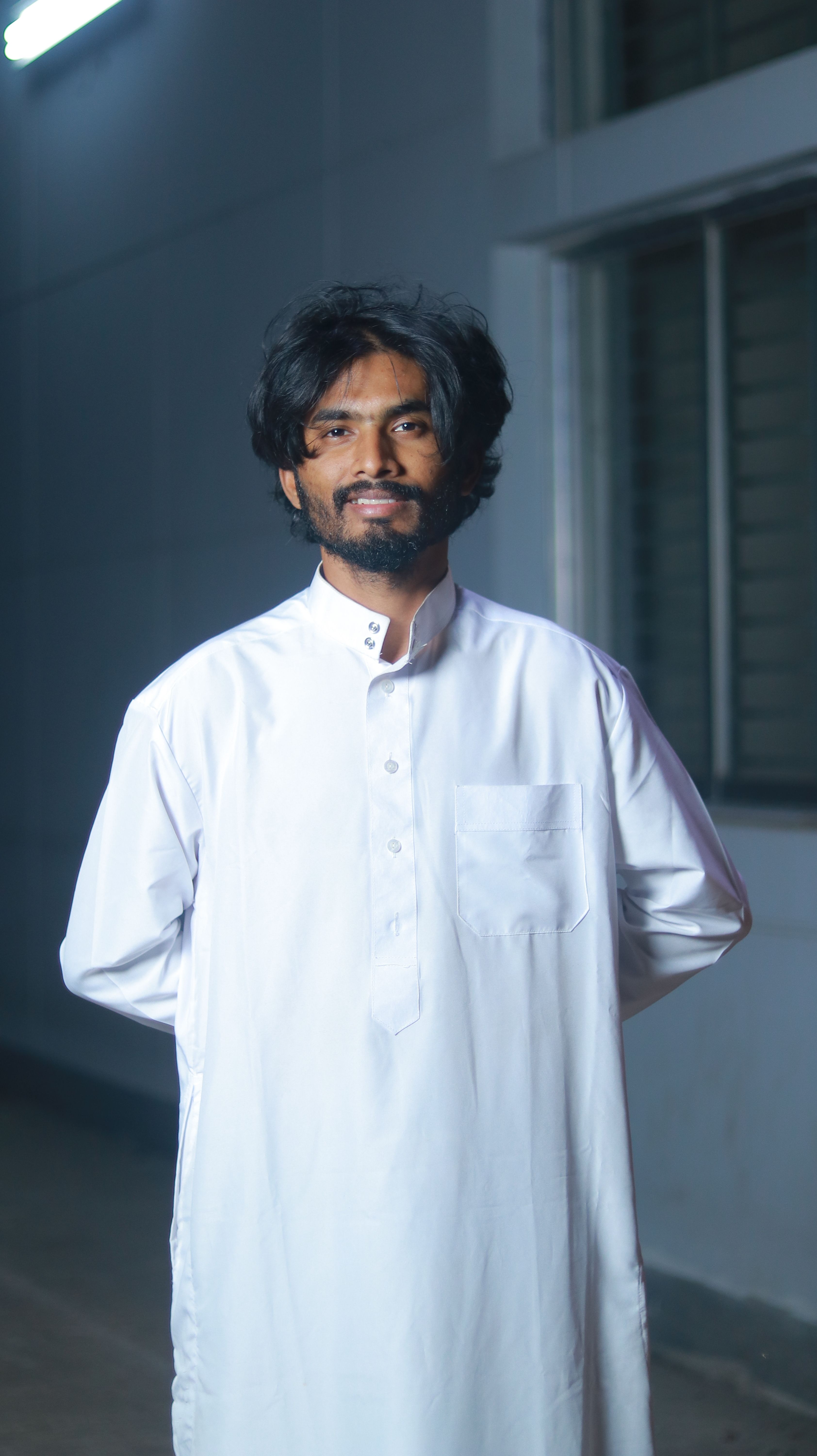 A man standing indoors, wearing traditional white clothing, smiling confidently at the camera.