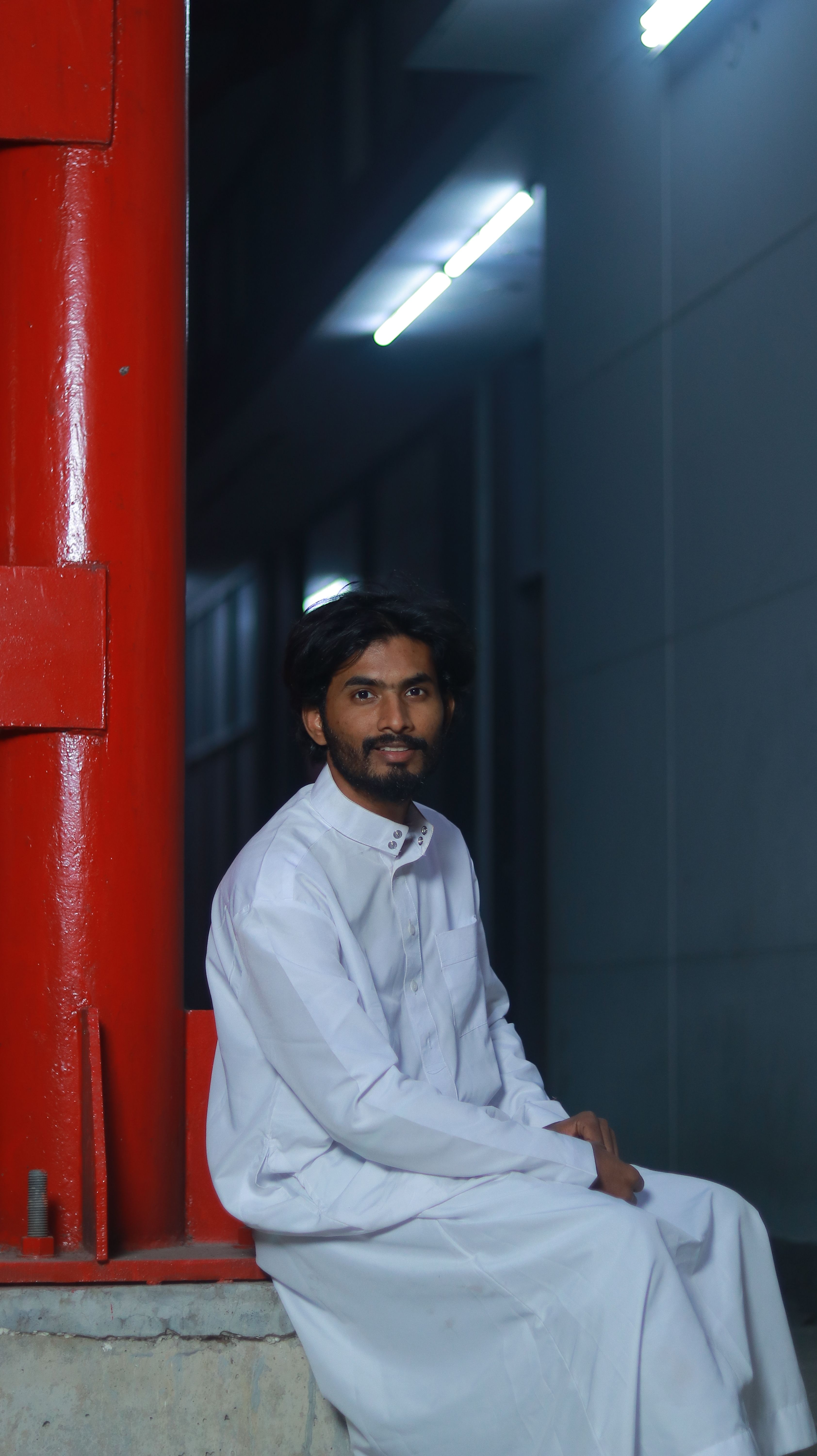 Portrait of a man in traditional attire sitting beside a red column at night.