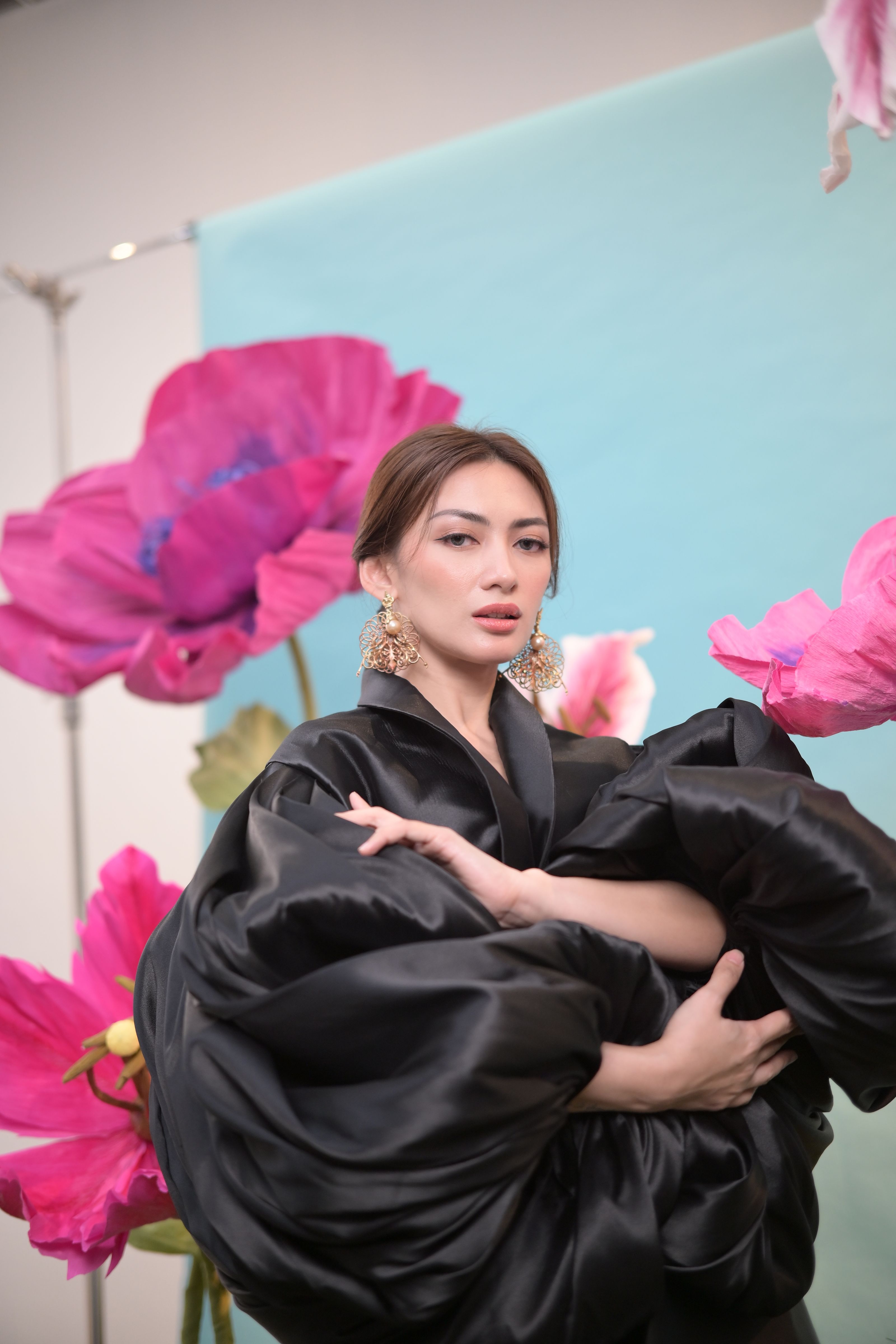 Mulher elegante de vestido preto posando entre grandes flores cor-de-rosa com um cenário sereno.