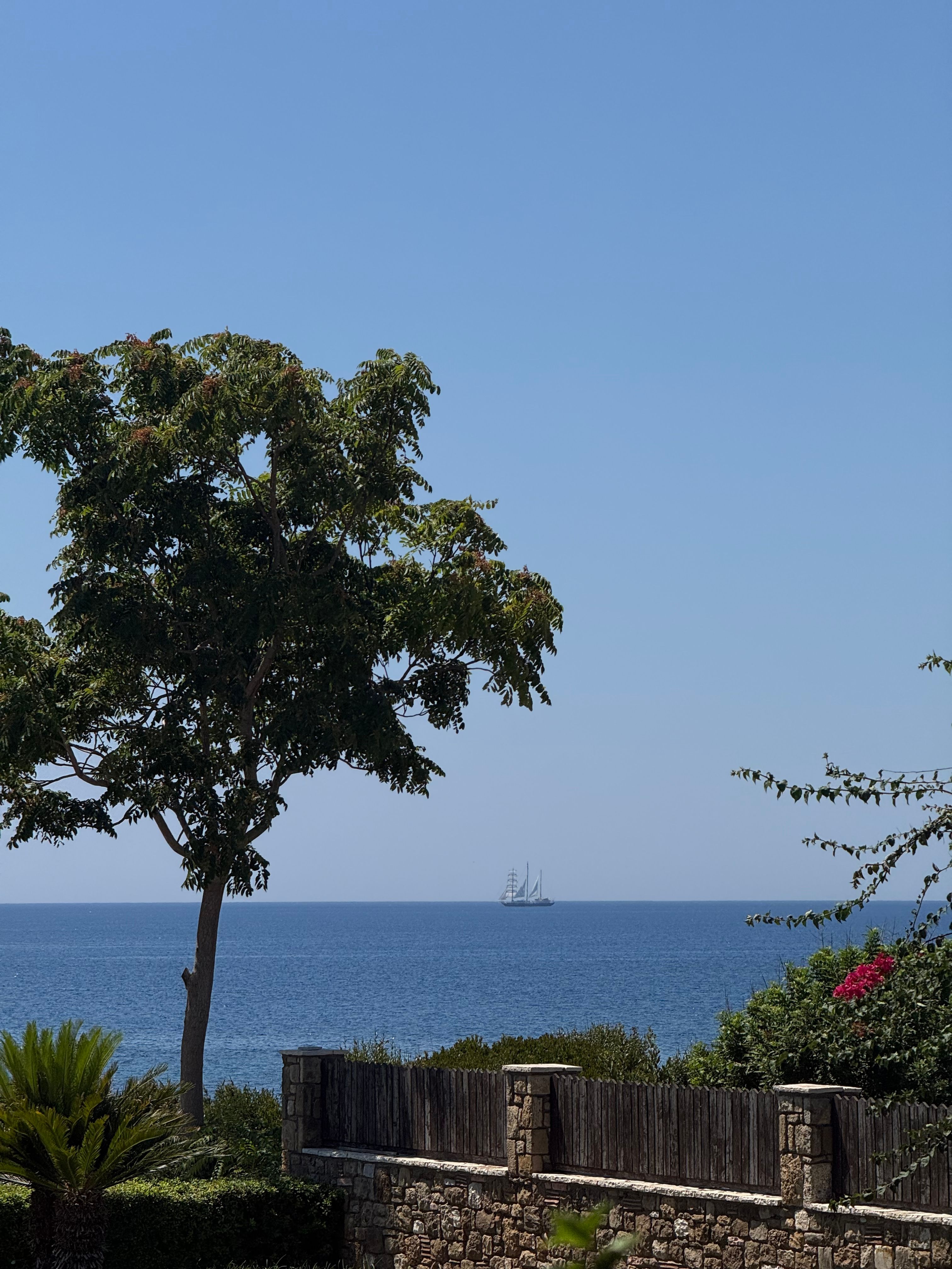 A tall sailing ship against a clear blue sky seen from a coastal garden in Greece.