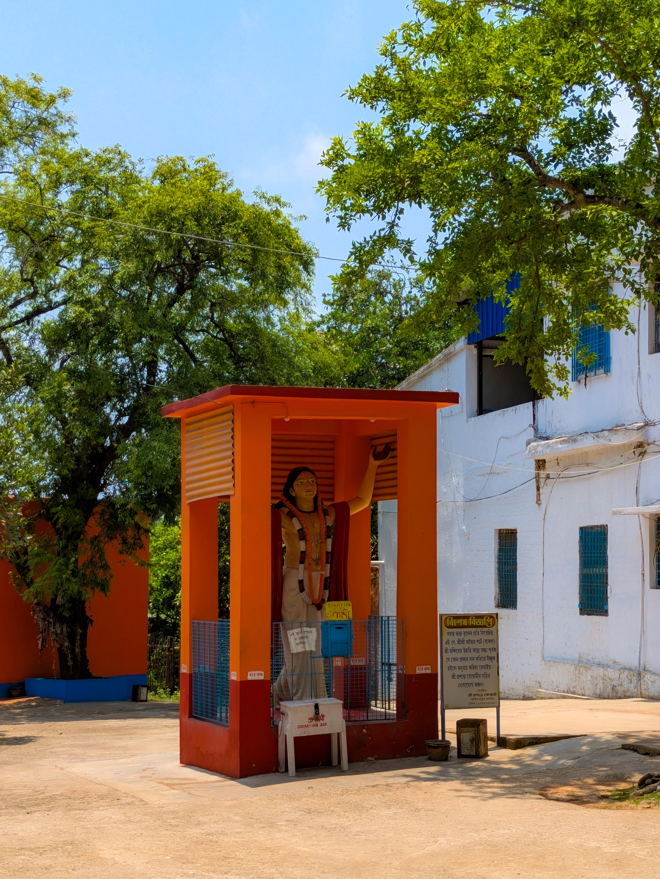 A vibrant shrine with a statue, surrounded by trees and adjacent to a white building outdoors.