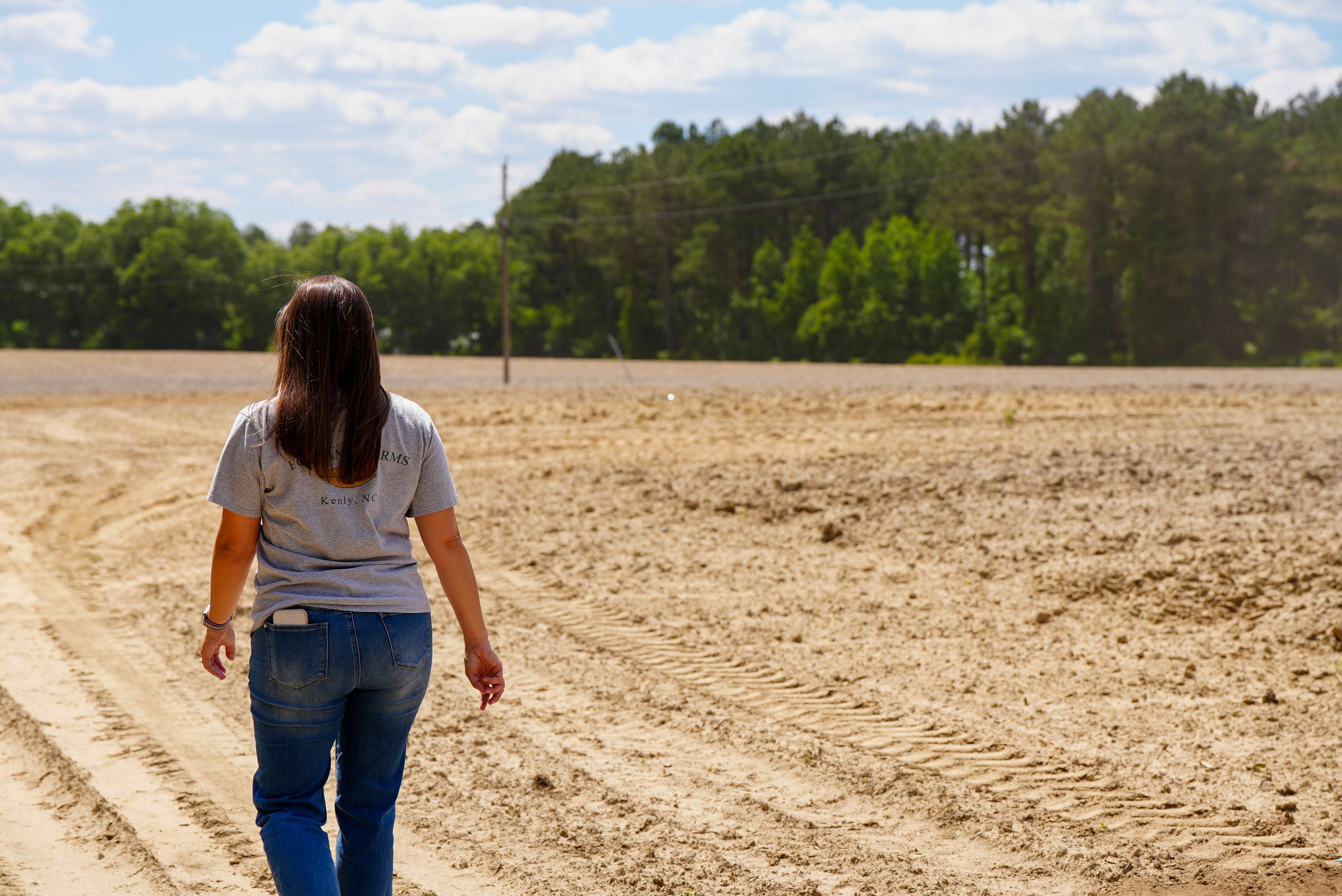 A woman walks down a rural dirt road, surrounded by fields and trees, under a bright sky.