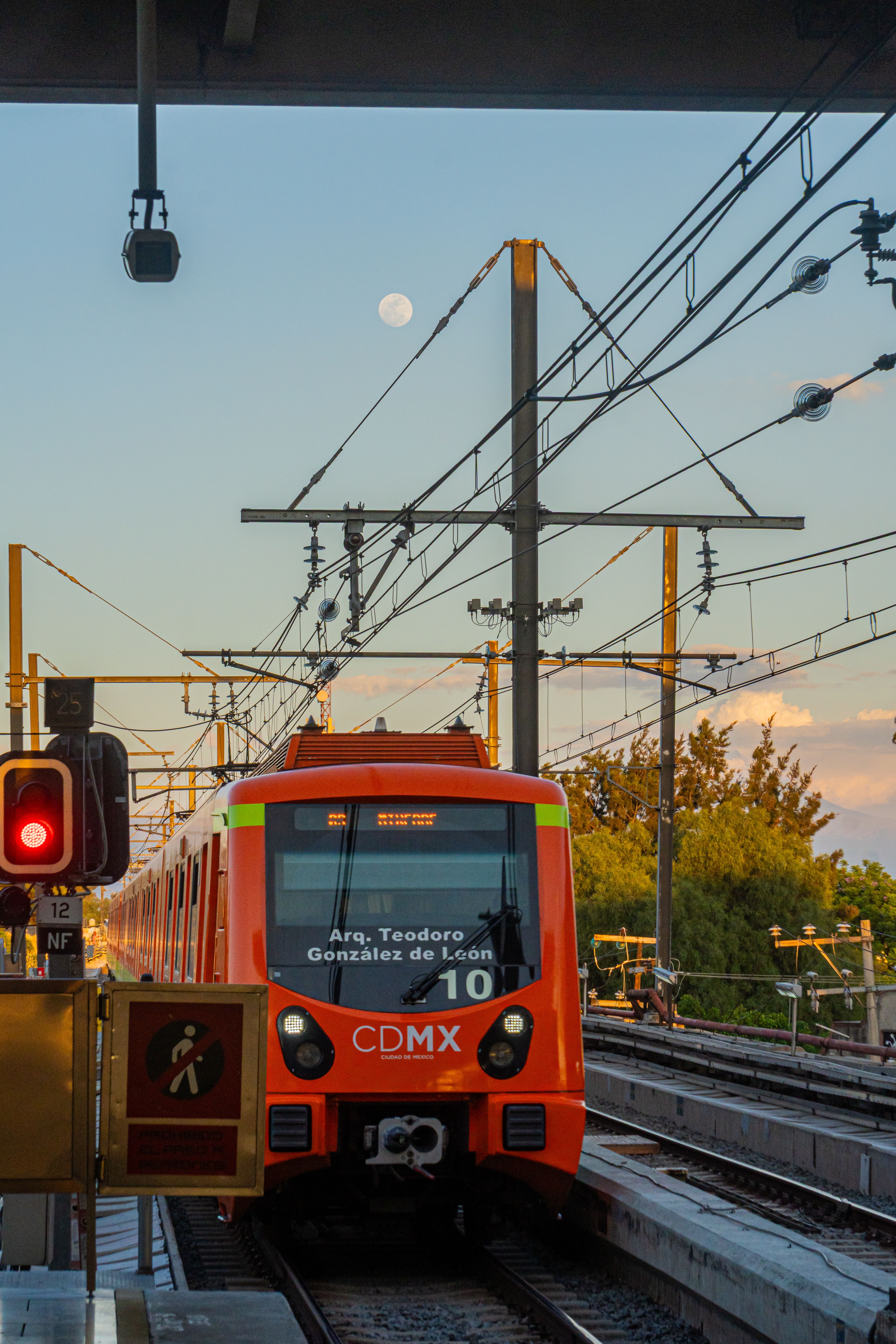 An orange CDMX metro train on the tracks at dusk with a clear moon in the sky.