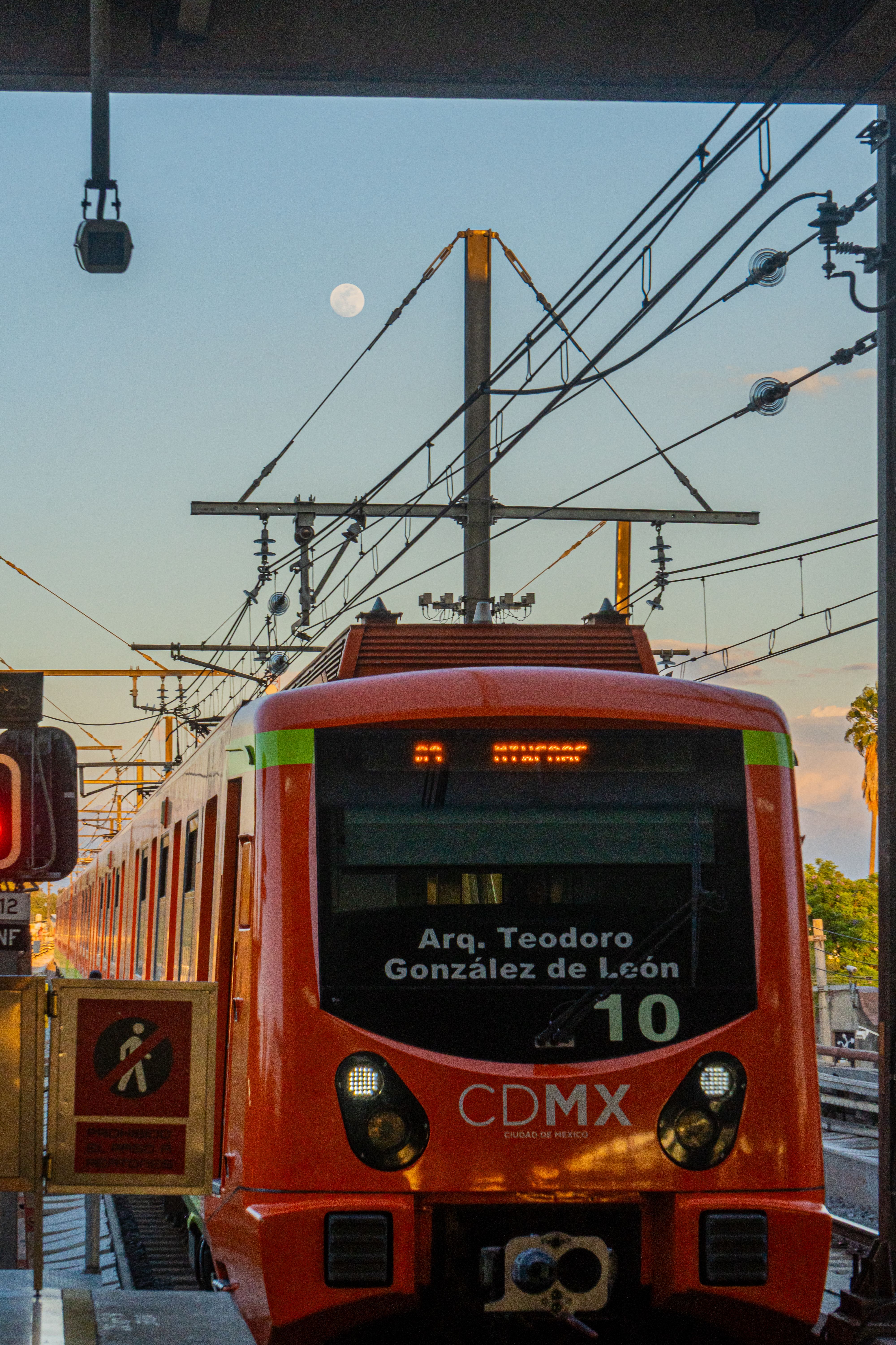 Vibrant orange metro train arriving at a station in Mexico City, highlighted by a visible moon in the evening sky.