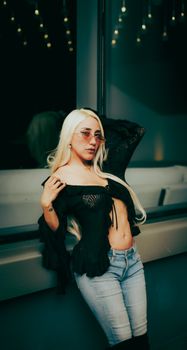 Confident young woman with sunglasses and fashionable attire posing indoors with ambient lighting.