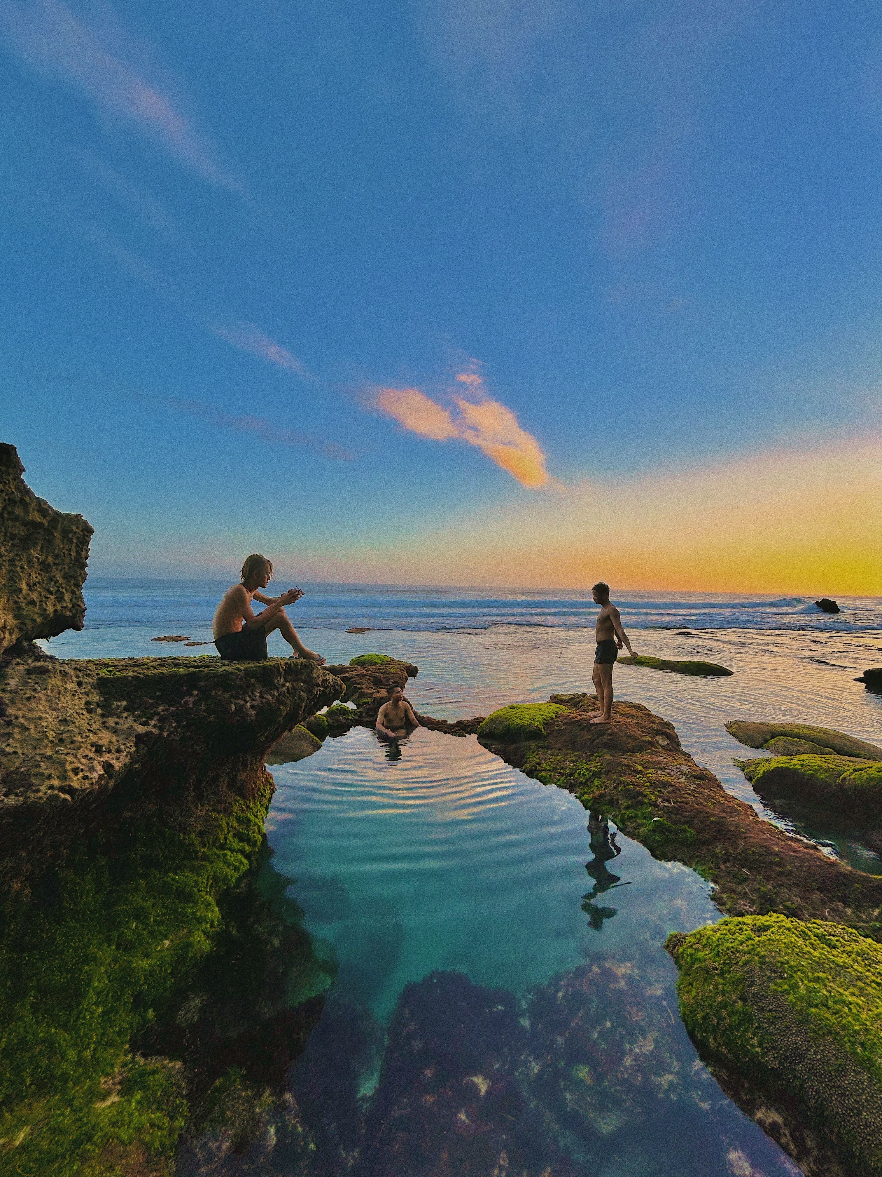 Three people enjoy a tranquil sunset at a rocky shoreline in Bali, Indonesia.
