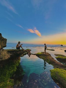 Three people enjoy a tranquil sunset at a rocky shoreline in Bali, Indonesia.