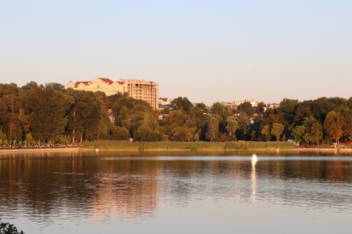 Free A peaceful lakeside scene with buildings and lush greenery illuminated by the warm glow of sunset. Stock Photo