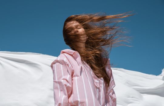 A woman in a striped pink shirt with her hair blown by the wind against a clear blue sky.