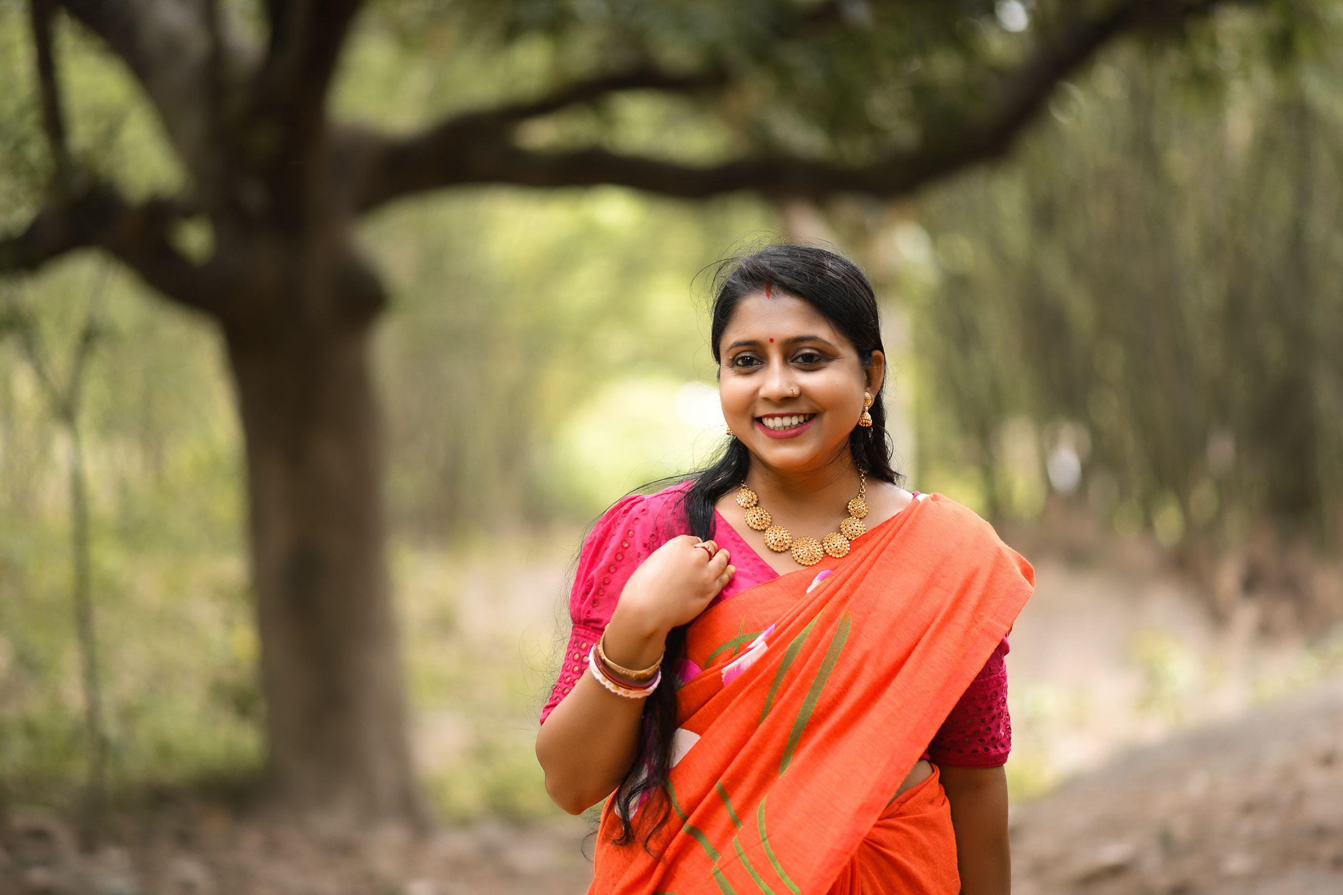 A smiling woman in an orange saree enjoying a day outdoors surrounded by trees.