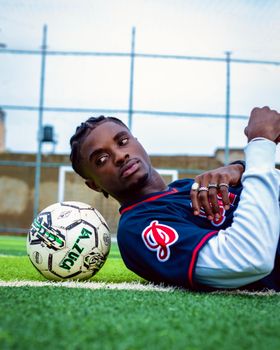 A young man lies on a soccer field in Kano, Nigeria, with a ball, showing casual confidence and style.