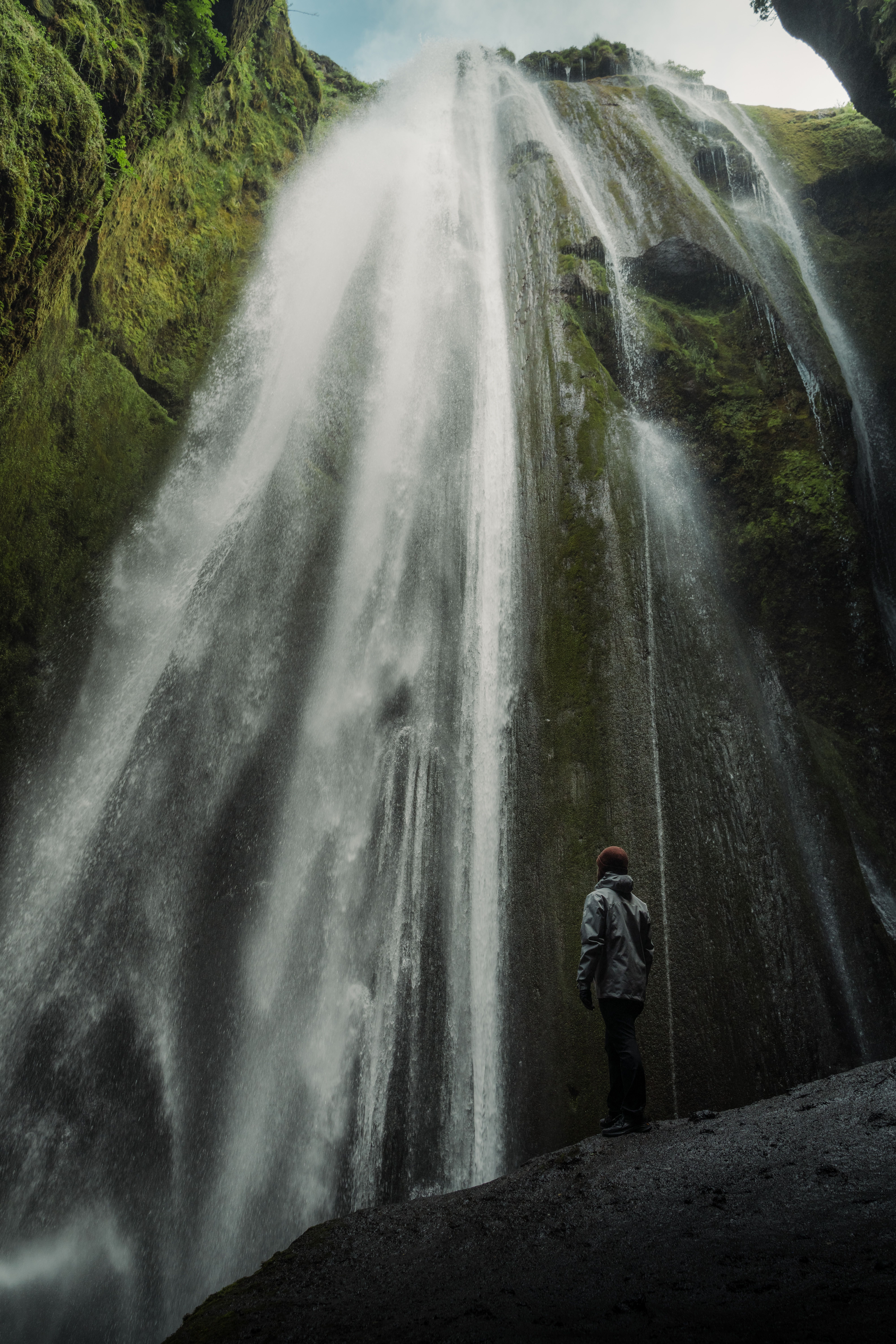 A lone traveler stands awe-inspired before a towering waterfall in a lush canyon.