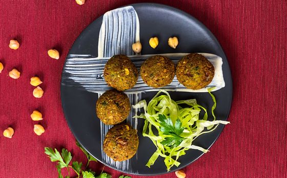 Five green-flecked falafel on tahini sauce with shredded lettuce and parsley on a dark plate