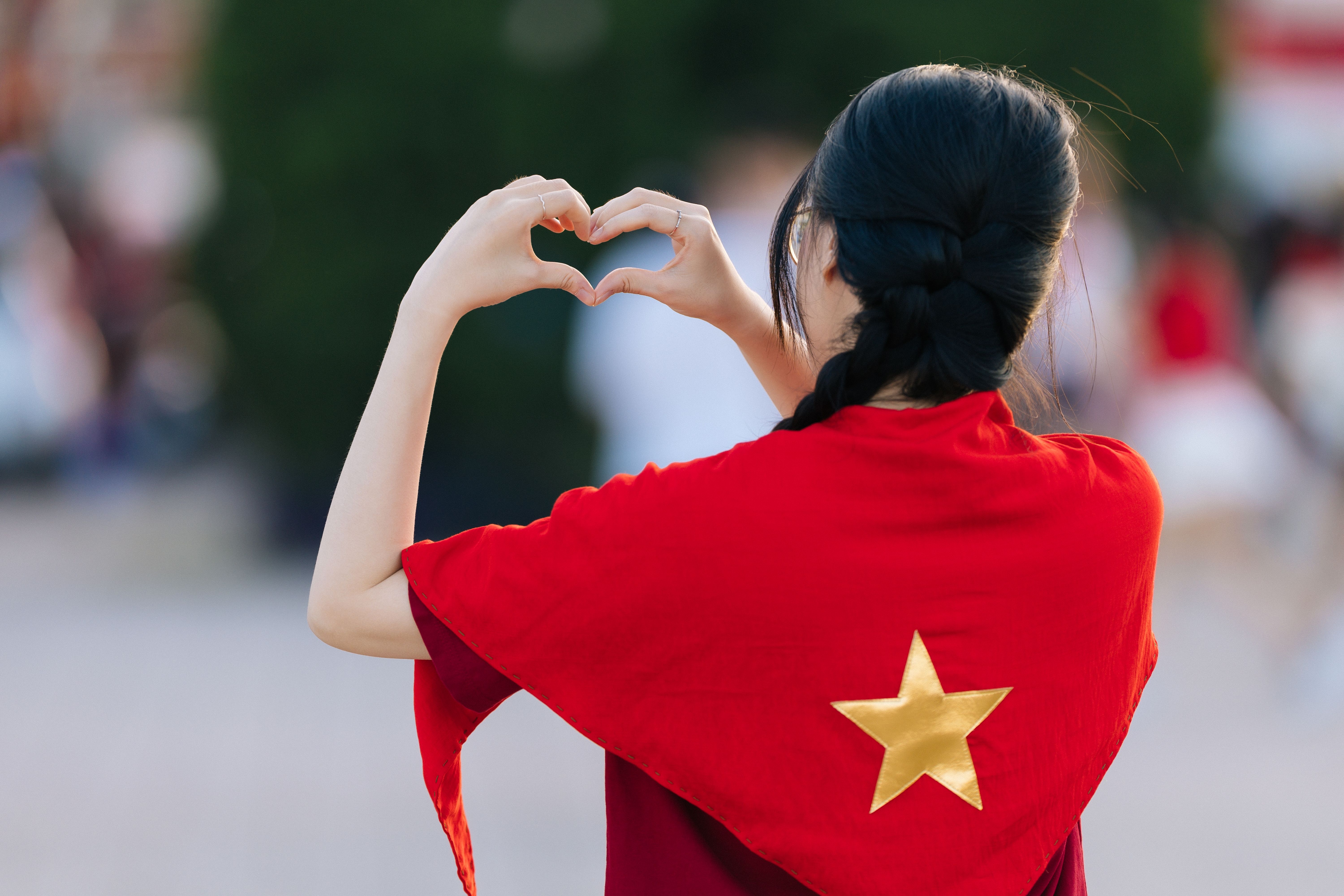Woman making heart shape with hands, draped in Vietnamese flag outdoors.