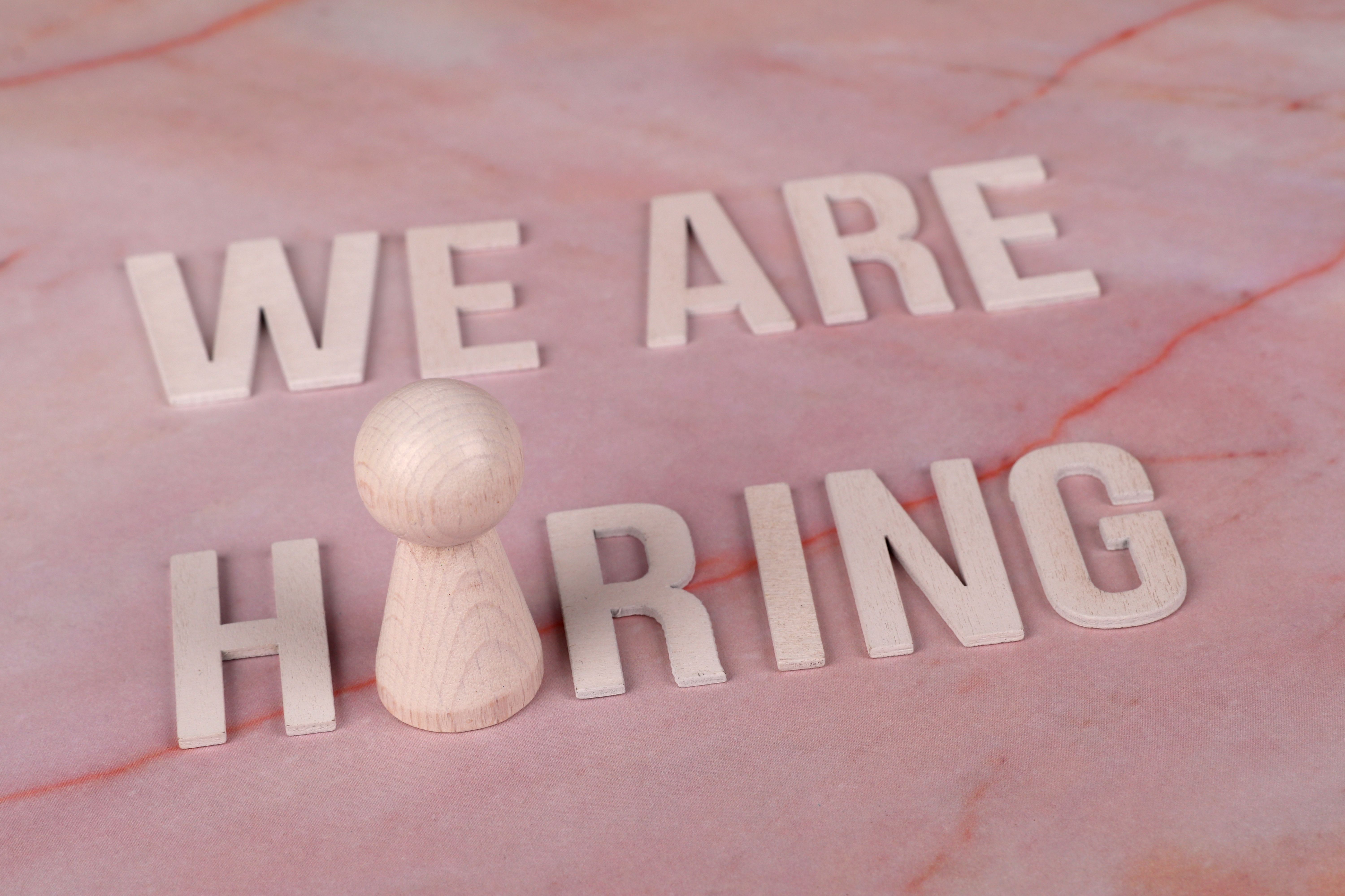 ピンクの大理石の表面に、ポーンの駒が置かれた状態で、「We Are Hiring（採用募集中）」と書かれた木製の文字。