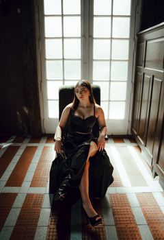 A woman in a long black dress sits gracefully in an elegant indoor setting with natural light.