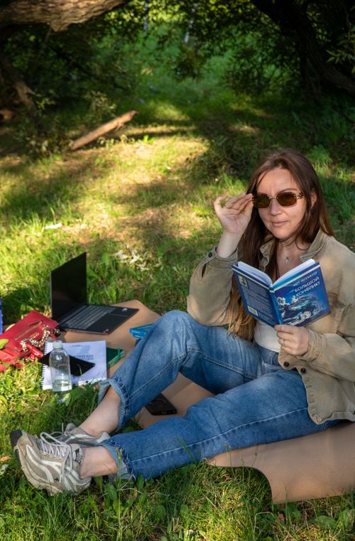 Free Woman enjoys a sunny day reading outdoors with a laptop and book in a park setting. Stock Photo