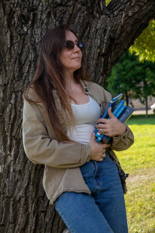 Free Young woman in casual attire leaning against a tree outdoors, holding books. Stock Photo