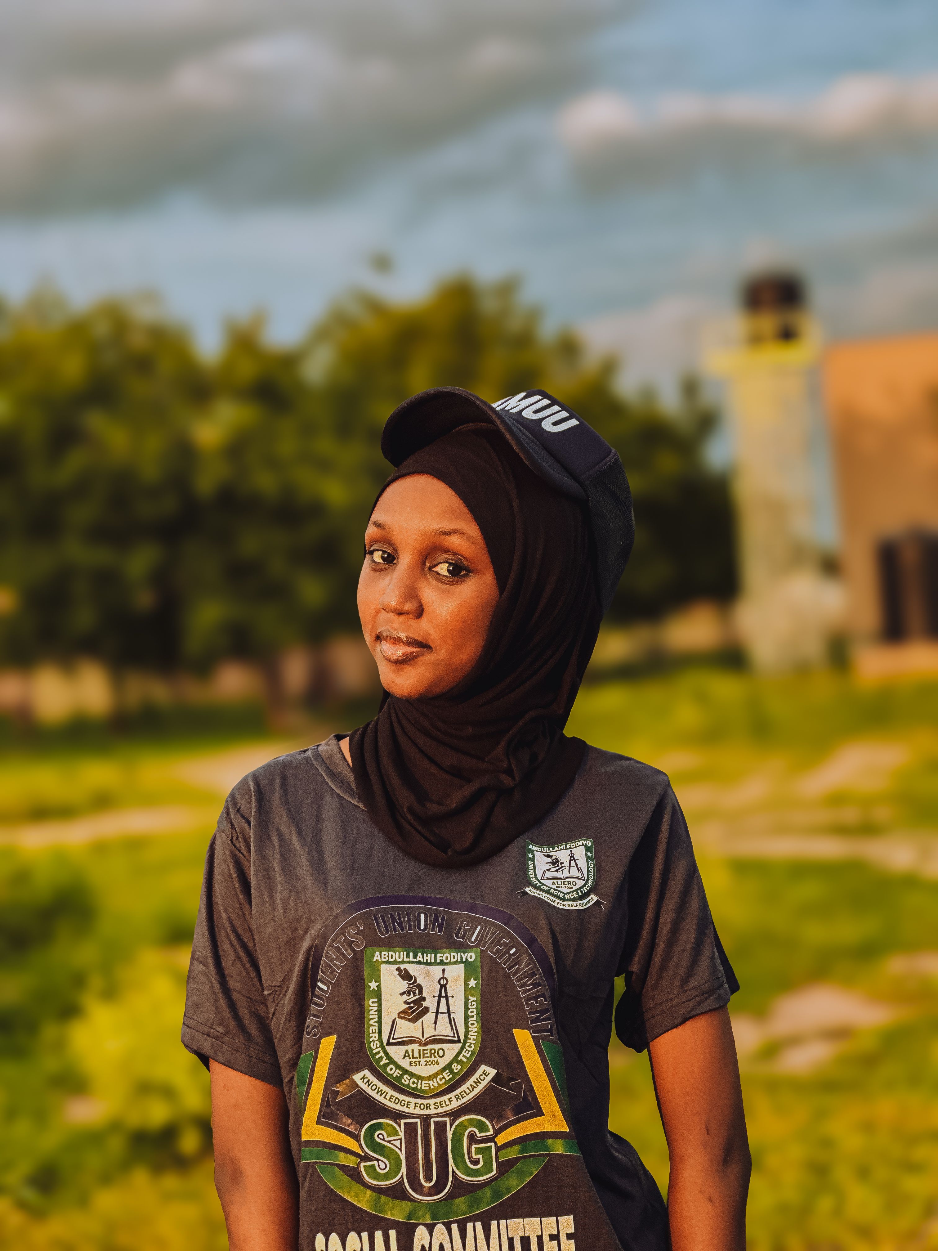 Young woman wearing cap and hijab standing outdoors on a sunny day.