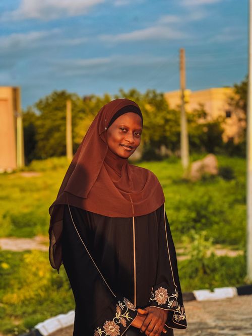 Free A woman in a brown hijab smiles warmly under the sunset light in a green outdoor setting. Stock Photo