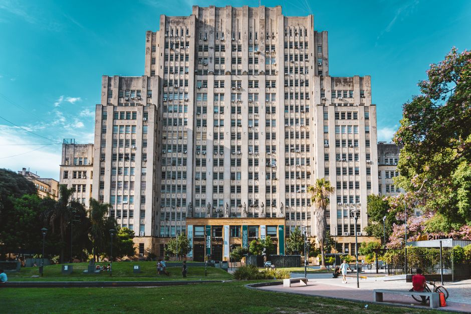 Un edificio universitario histórico que exhibe arquitectura art déco con un frondoso parque en primer plano.