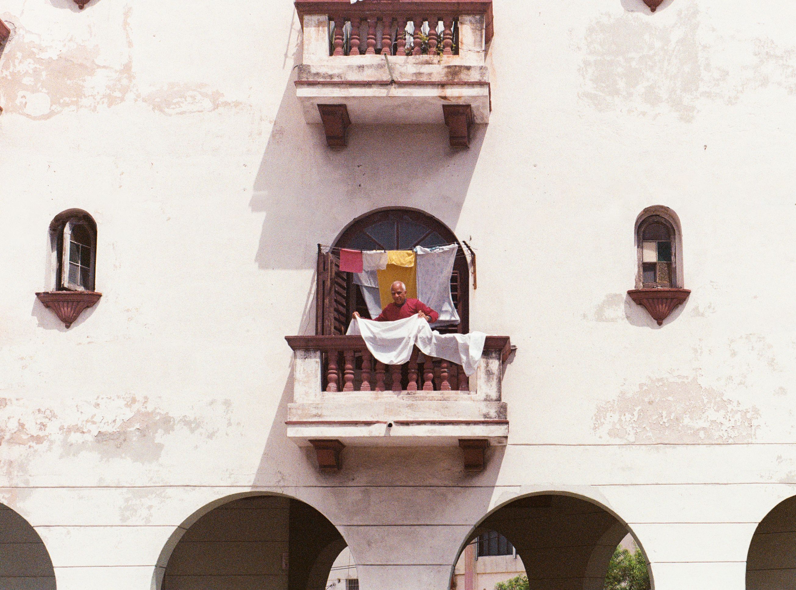 A man stands on a balcony with hanging laundry and colorful prayer flags, against an aged building facade.