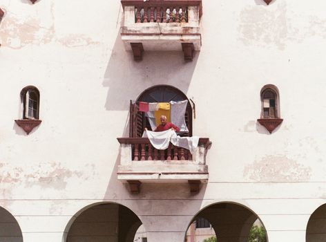 A man stands on a balcony with hanging laundry and colorful prayer flags, against an aged building facade.