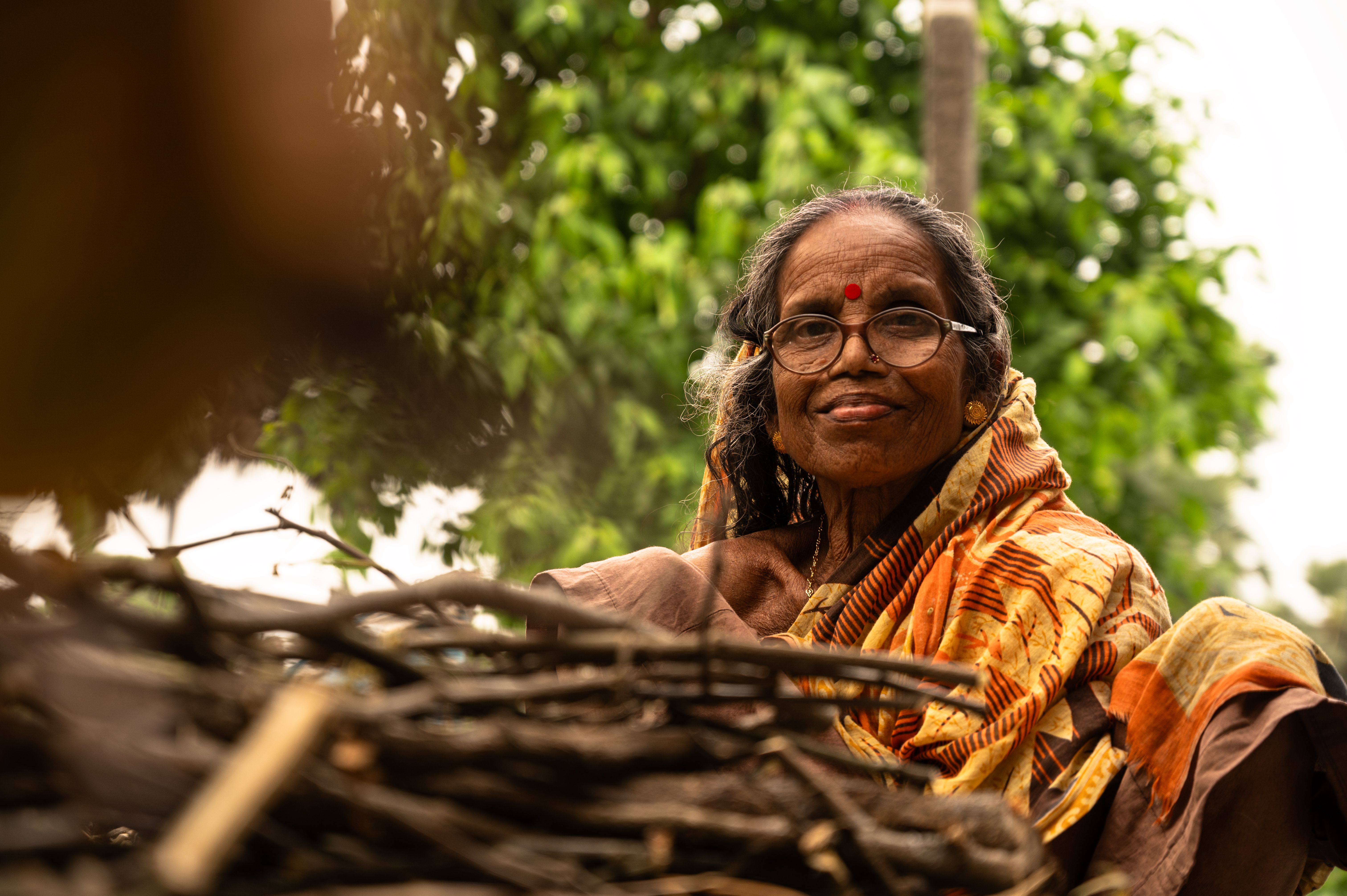 Elderly woman in sari smiling near stacked firewood, outdoors.