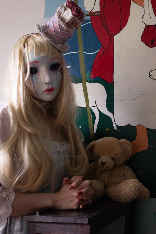 Free Whimsical masked figure with a doll-like appearance poses indoors with a teddy bear, evoking mystery. Stock Photo