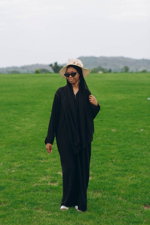 Free A woman in a black kaftan with sunglasses and hat walks in a sunny green park. Stock Photo