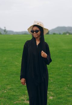 A fashionable woman wearing sunglasses and a hijab enjoys a sunny day in a lush green park.