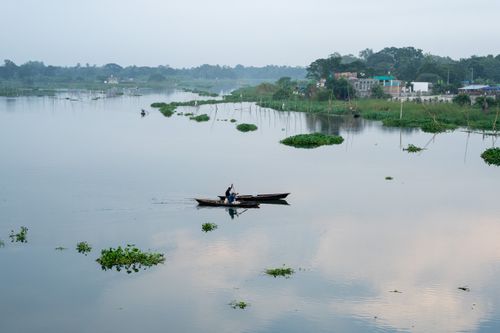 Free A tranquil river landscape with a small boat carrying fishermen at dawn, surrounded by greenery. Stock Photo