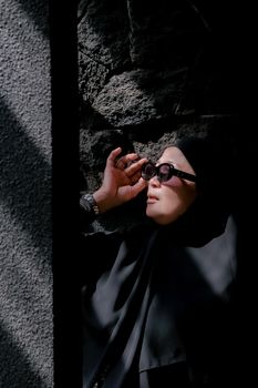 An adult in sunglasses adjusts them under dramatic lighting against a textured wall.