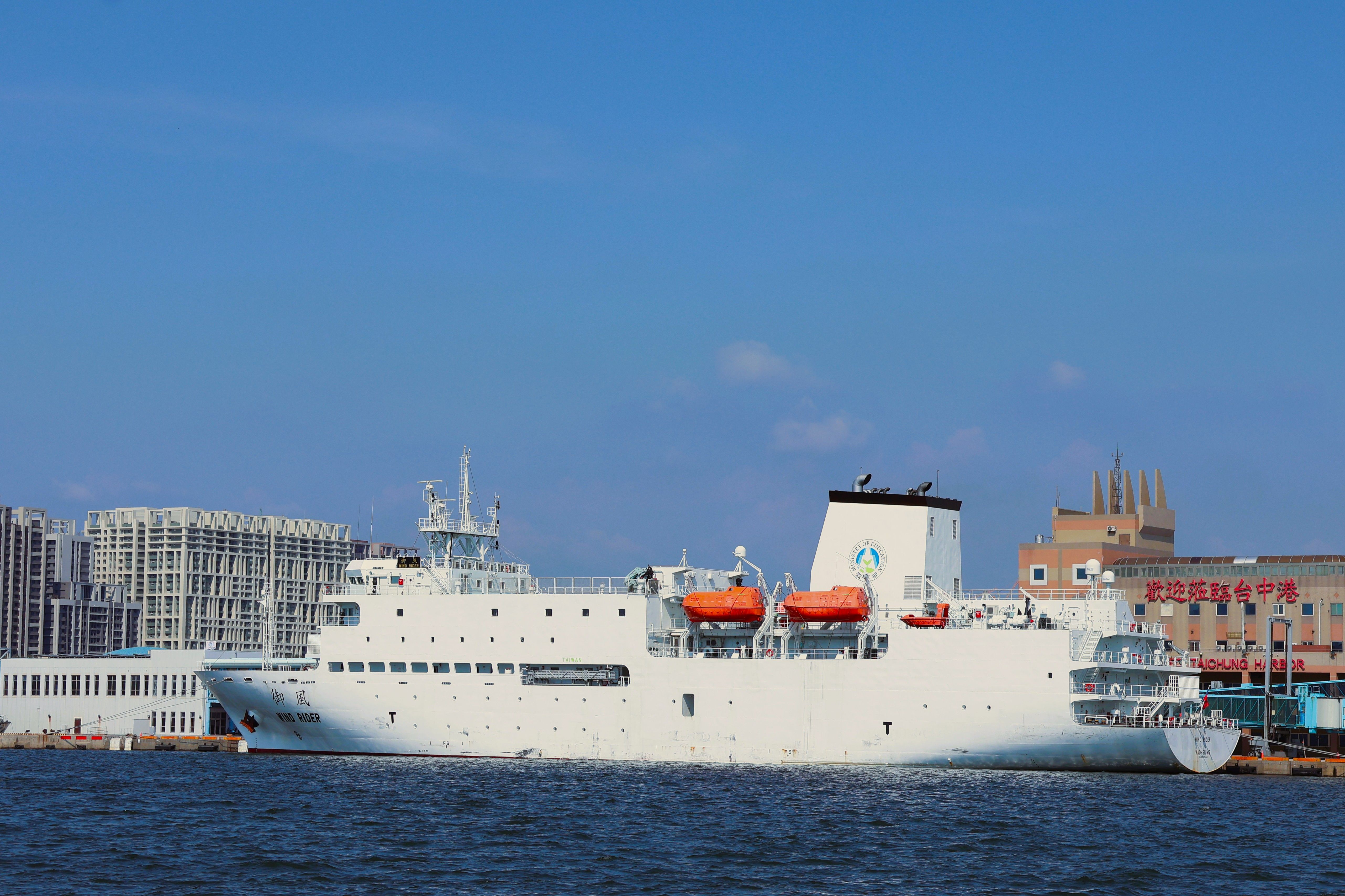 A large white ship with orange lifeboats docked at Taichung Harbor, under a clear blue sky.