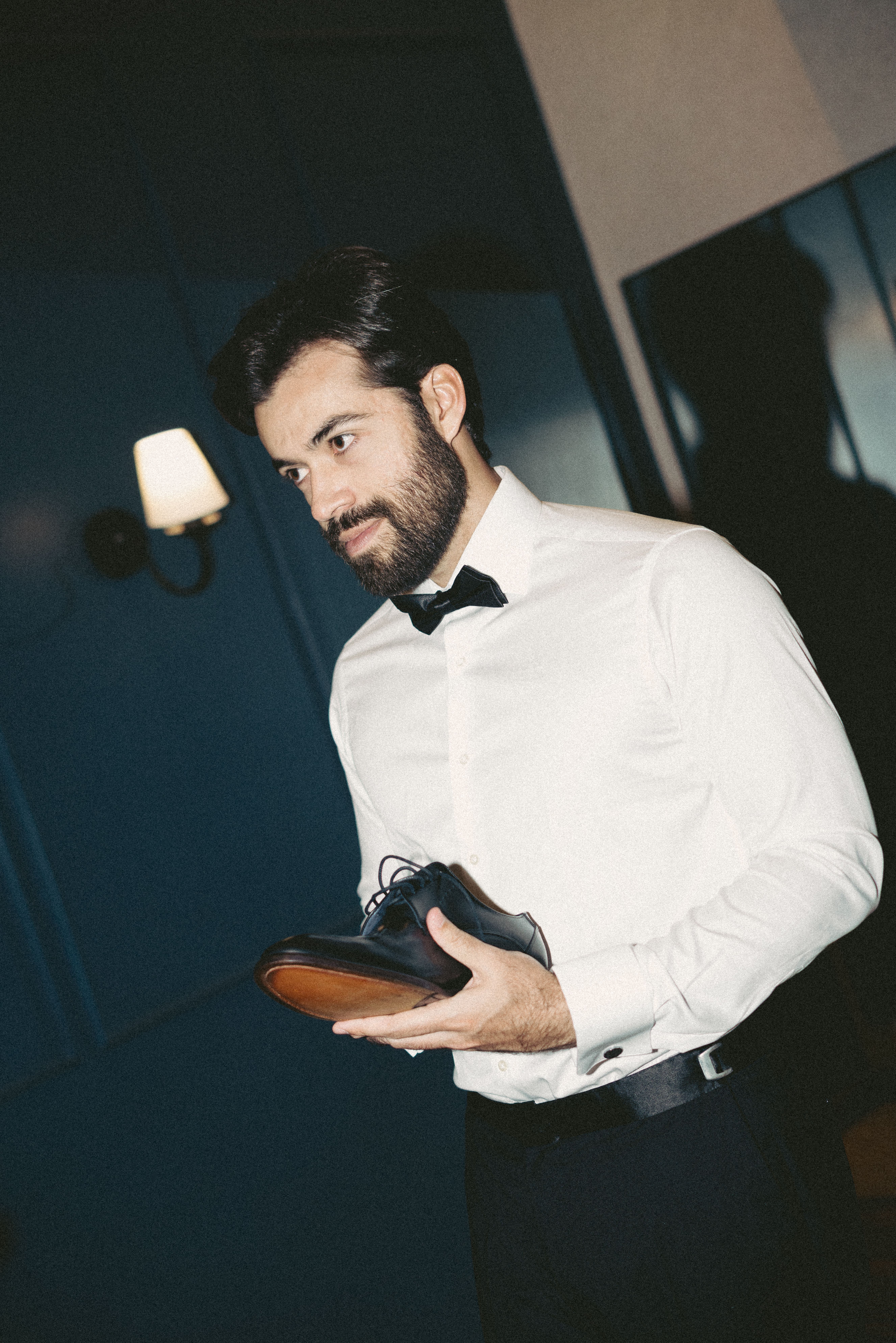 A well-dressed man in a white shirt and bow tie holding a black shoe, ready for a formal occasion.