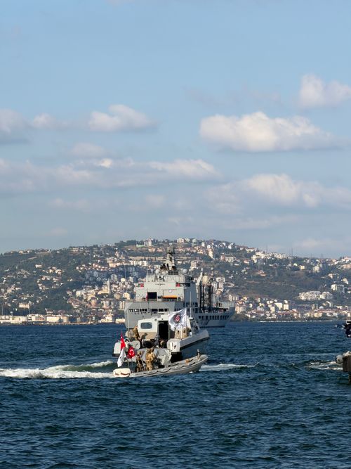 Free A naval ship approaches a seaside town with hills and buildings under a partly cloudy sky. Stock Photo