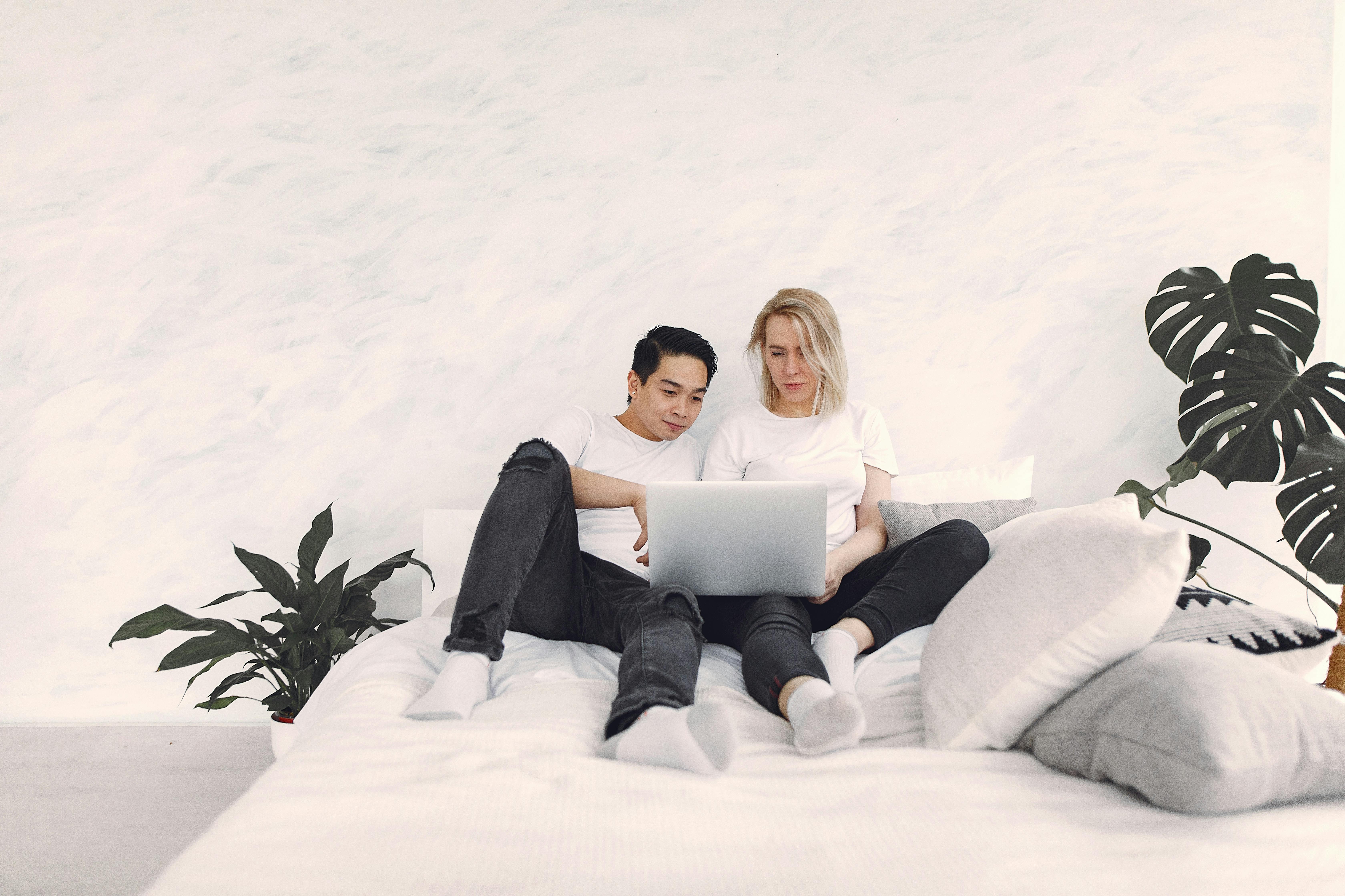 A couple lounging on a bed, sharing a laptop in a bright, minimalist bedroom setting.
