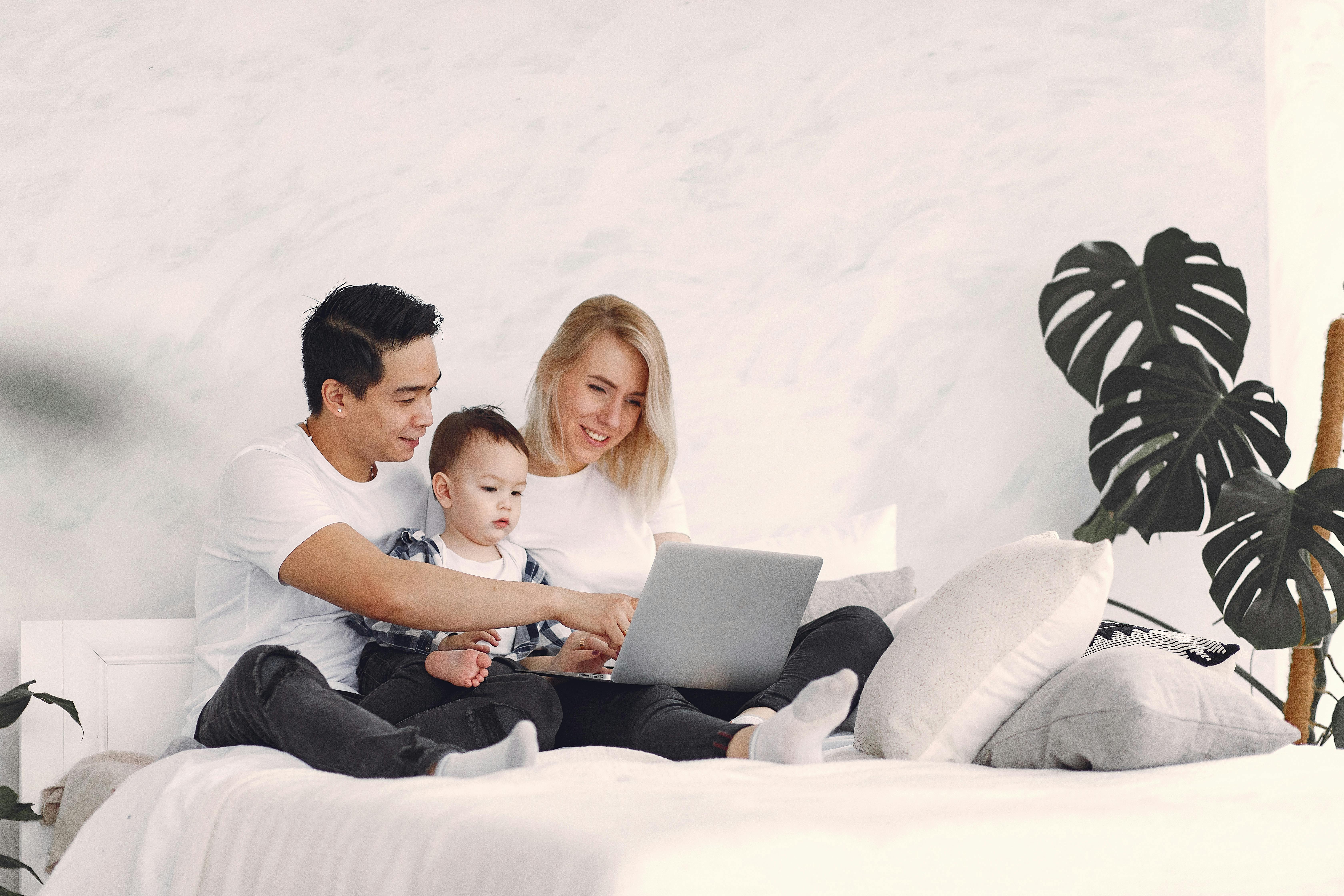 A joyful family with a child enjoying time together on a bed, using a laptop indoors.