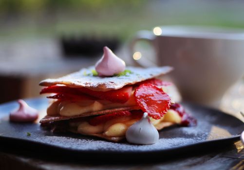 Close-up of a strawberry mille-feuille dessert with meringue and cup in background.