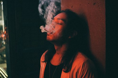 Free A contemplative man with long hair is smoking indoors at night, casting a dramatic shadow. Stock Photo