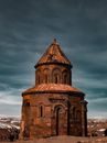 Amazing view of aged medieval chapel exterior with brown walls and dome against dark cloudy sky in ruined city of Ani in Armenia