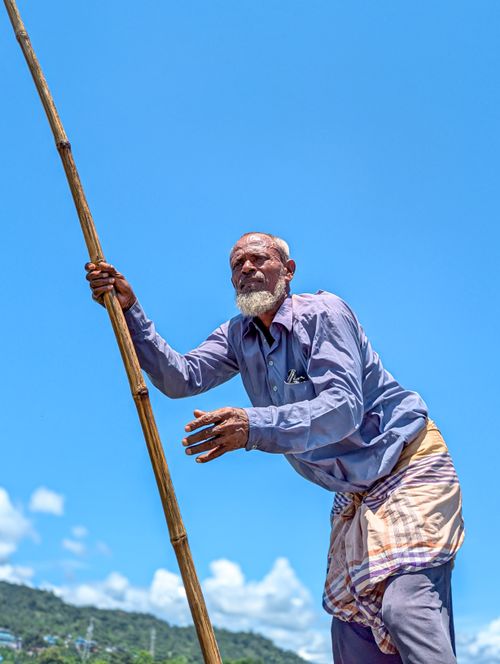 Free An elderly boatman uses a bamboo pole to navigate under a clear blue sky in Sylhet, Bangladesh. Stock Photo